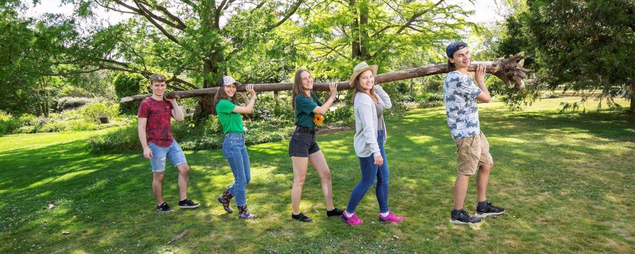 junge Menschen tragen einen Baumstamm über eine Wiese