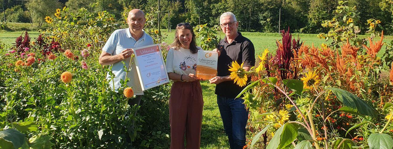 Bürgermeister Michael Krischke, Sonja Broser, BP-Direktor Andreas Weiß mit Urkunde im Blumenfeld