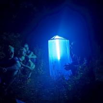 Nachts sitzen Menschen im Freien um ein leuchtendes, würfelförmiges Objekt mit intensivem blauem Licht. Umgebung ist von Bäumen umgeben